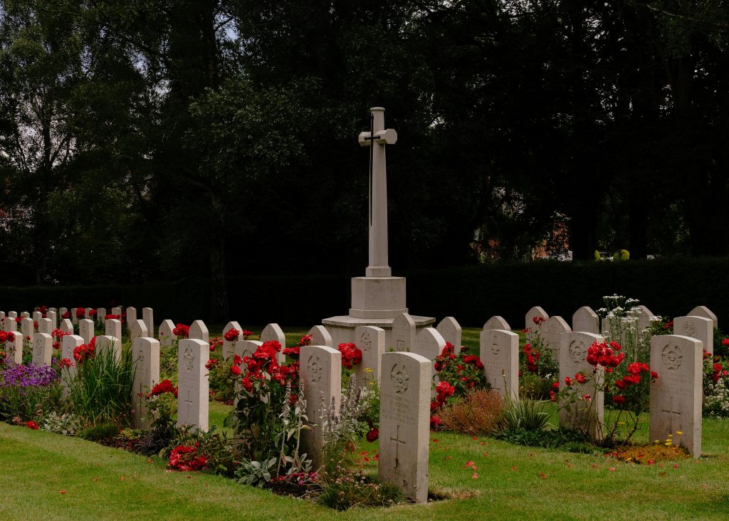 Peaceful cemetery scene with gravestones, cross, and vibrant flowers under mature trees.